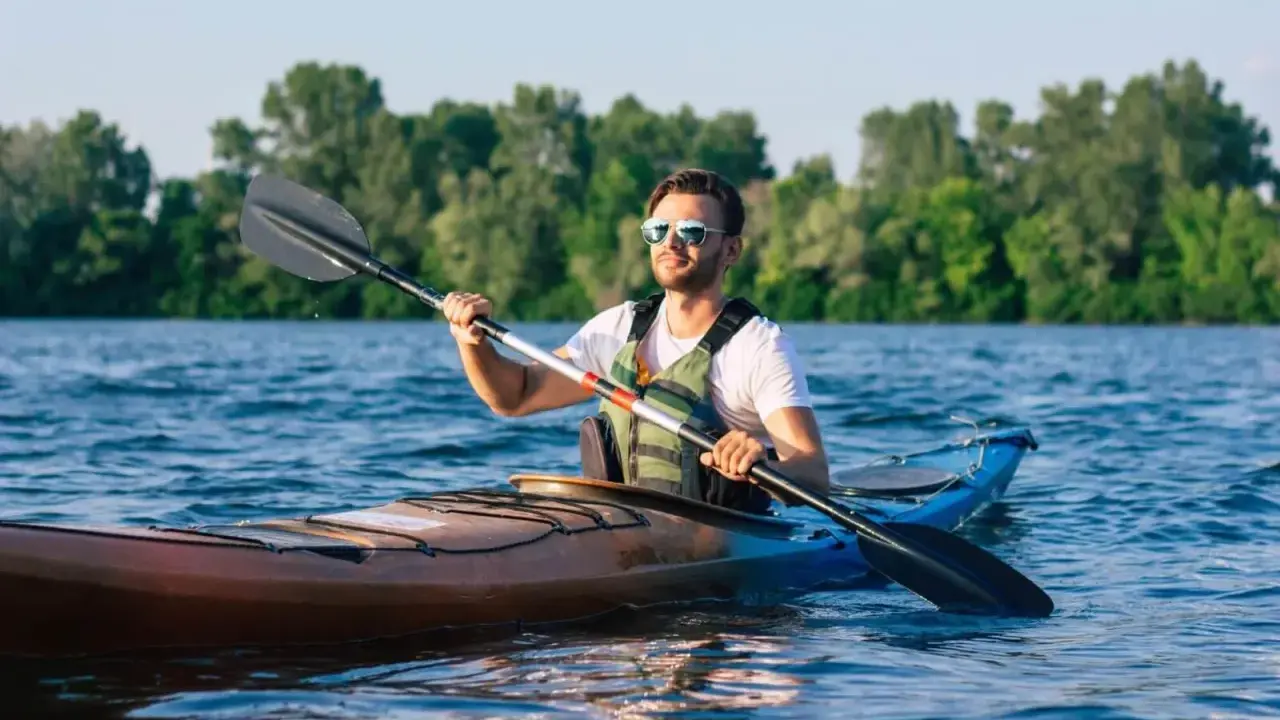 Mężczyzna w okularach przeciwsłonecznych i kamizelce ratunkowej wiosłuje kajakiem po spokojnej rzece. Idealne miejsce na najlepsze spływy kajakowe.