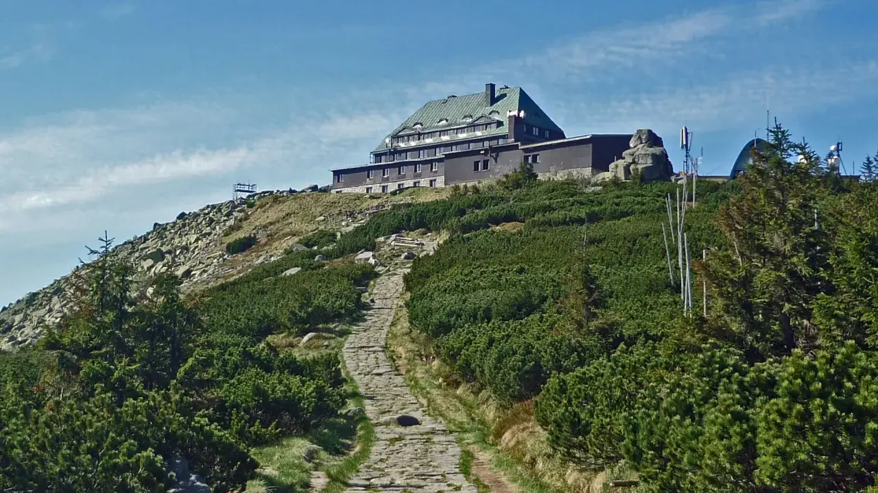 Kamienista ścieżka prowadzi na szczyt, gdzie widać schronisko. Wokół bujna roślinność i skaliste zbocza.