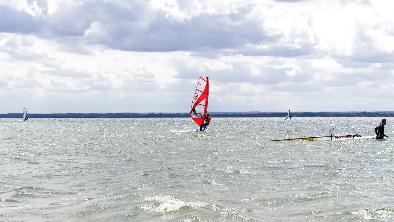 Czerwony żagiel do windsurfingu na wodzie, surfer w akcji. W tle inni windsurferzy i chmury.