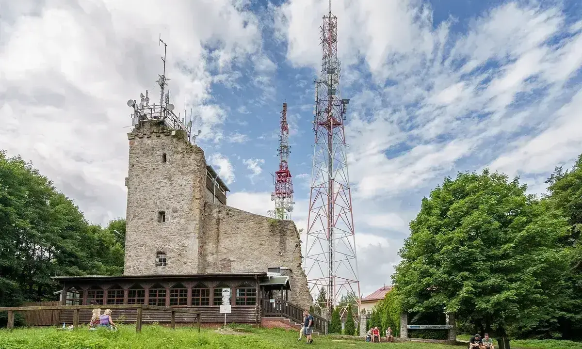 Kamienna wieża i wysokie maszty telekomunikacyjne na szczycie Chełmiec. Ludzie odpoczywają na trawie.