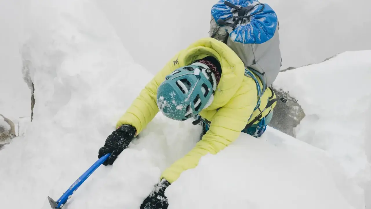 Alpinista w żółtej kurtce i niebieskim kasku wspina się po śnieżnej grani, używając czekana do hamowania.
