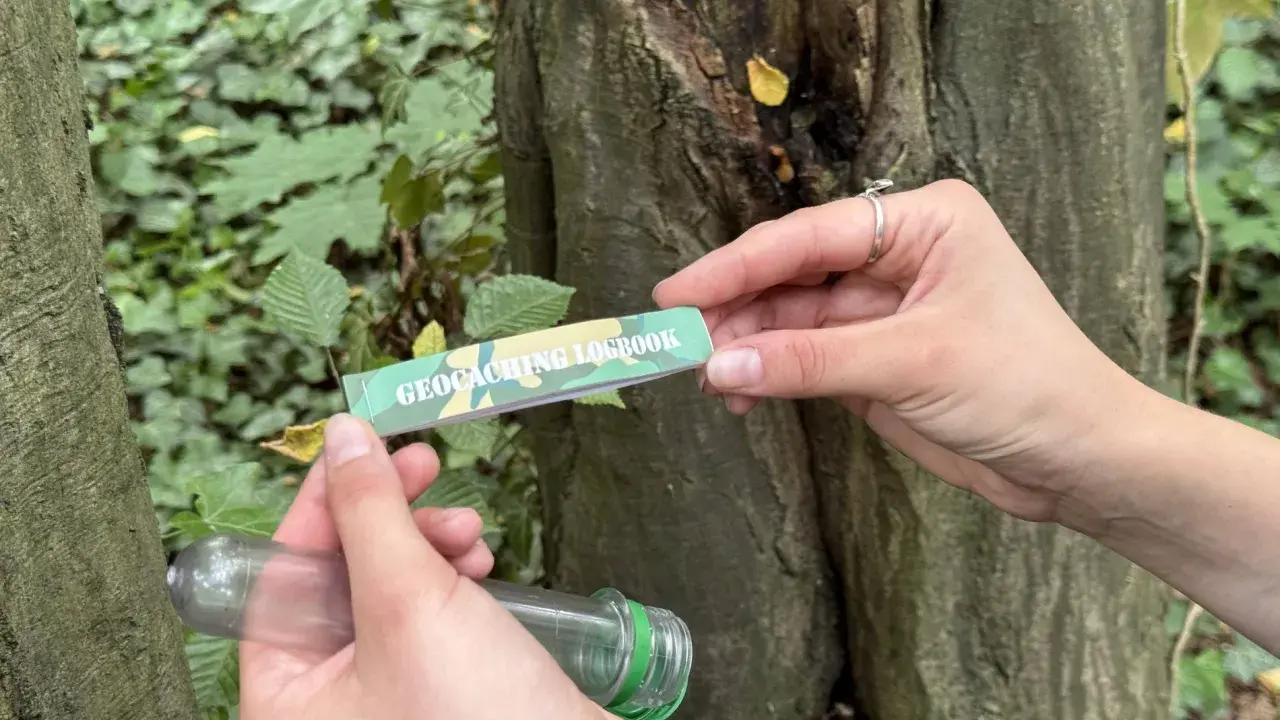 Ręce trzymają pojemnik i zeszyt z napisem "GEOCACHING LOGBOOK". To wyjaśnia, geocaching co to jest: poszukiwanie ukrytych skarbów.
