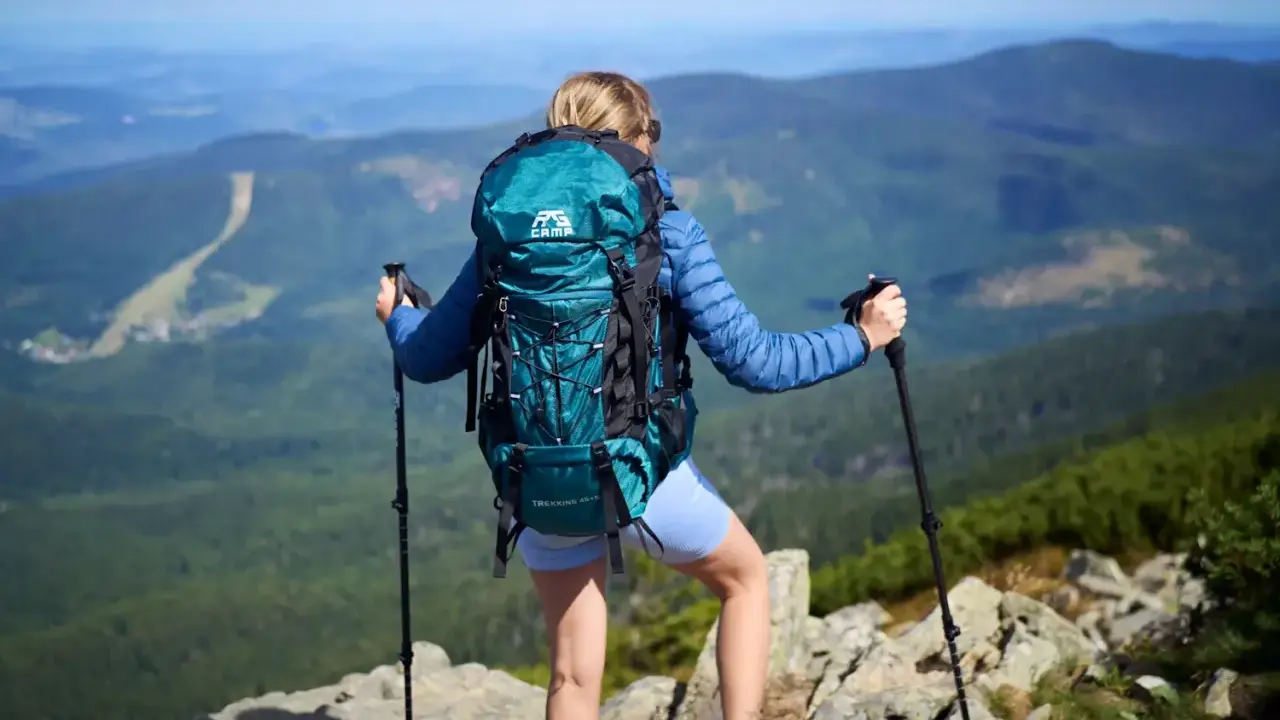 Kobieta z turkusowym plecakiem w góry, z kijkami trekkingowymi, podziwia panoramę.