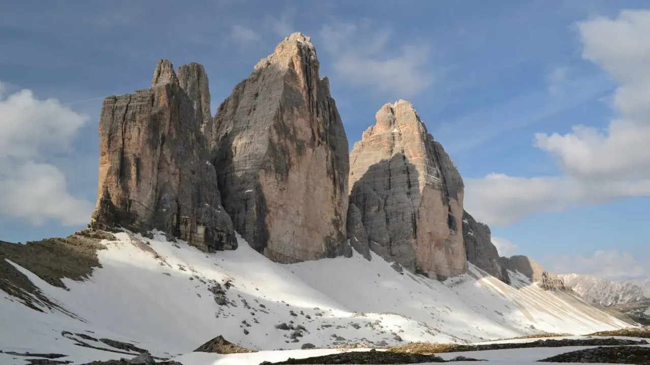 Majestatyczne **Tre Cime** w Dolomitach, ośnieżone zbocza i błękitne niebo z chmurami.