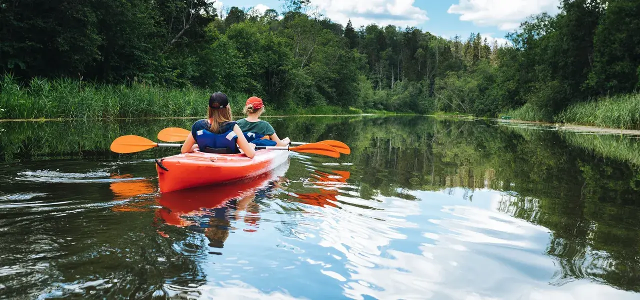 Dwie osoby płyną pomarańczowym kajakiem po spokojnej rzece. Wokół bujna zieleń drzew i trzcin. Idealny spływ kajakowy Mazury.