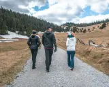 Trzy osoby idące ścieżką w górach. W tle las i pagórkowaty krajobraz.