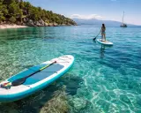 Sup chorwacja! Dwie deski SUP unoszą się na krystalicznie czystej wodzie. Jedna z nich, bliżej, ma wiosło. Na drugiej, dalej, stoi kobieta. W tle widać plażę i góry.
