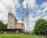 Kamienna wieża i wysokie maszty telekomunikacyjne na szczycie Chełmiec. Ludzie odpoczywają na trawie.