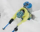 Alpinista w żółtej kurtce i niebieskim kasku wspina się po śnieżnej grani, używając czekana do hamowania.