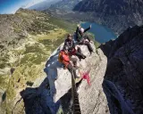 Trzech turystów siedzi na skale na grani wideł, machając do kamery. W tle jezioro i góry.