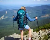 Kobieta z turkusowym plecakiem w góry, z kijkami trekkingowymi, podziwia panoramę.