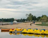 Żółte rowerki wodne i łódki czekają na plaży nad jeziorem blisko Warszawy. Para spaceruje po piasku pod słomianymi parasolami.