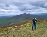 Kobieta z plecakiem na tle górskiego krajobrazu. W oddali widać pasma górskie, a na pierwszym planie trawiaste zbocze. To fragment Wielkiego Szlaku Beskidzkiego.