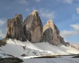 Majestatyczne **Tre Cime** w Dolomitach, ośnieżone zbocza i błękitne niebo z chmurami.