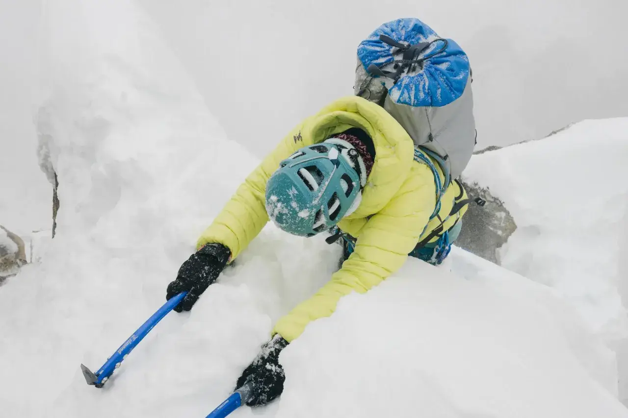 Alpinista w żółtej kurtce i niebieskim kasku wspina się po śnieżnej grani, używając czekana do hamowania.