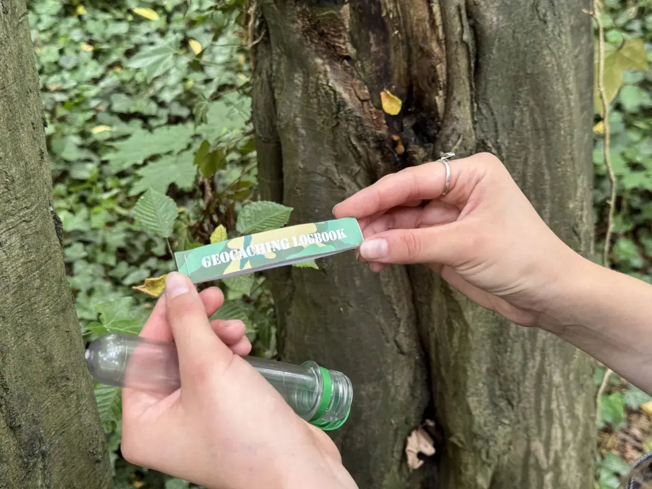Ręce trzymają pojemnik i zeszyt z napisem "GEOCACHING LOGBOOK". To wyjaśnia, geocaching co to jest: poszukiwanie ukrytych skarbów.