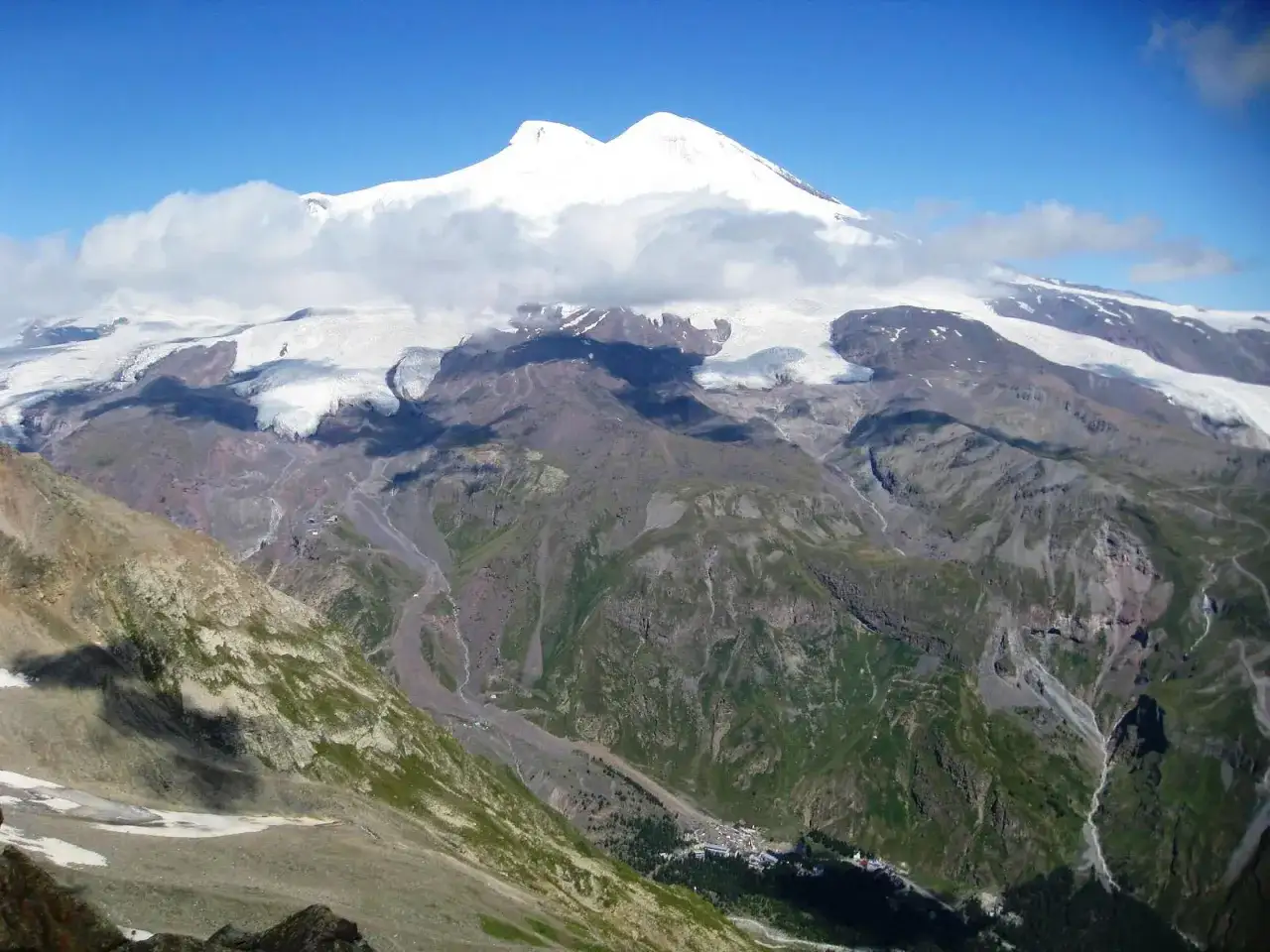 Elbrus, najwyższy szczyt Rosji, w chmurach. Lodowce spływają po zboczach, a w dolinie widać małą osadę.