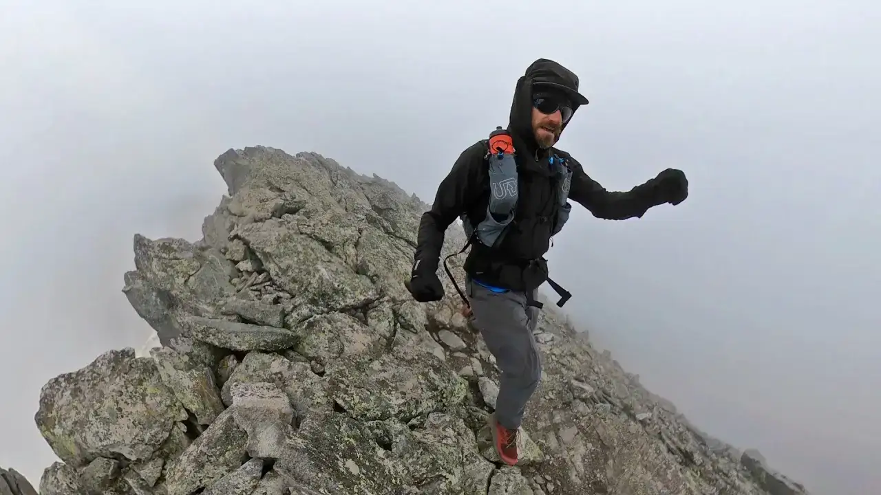 Mężczyzna w czapce i okularach przeciwsłonecznych uprawia speed hiking po skalistym, mglistym szczycie.