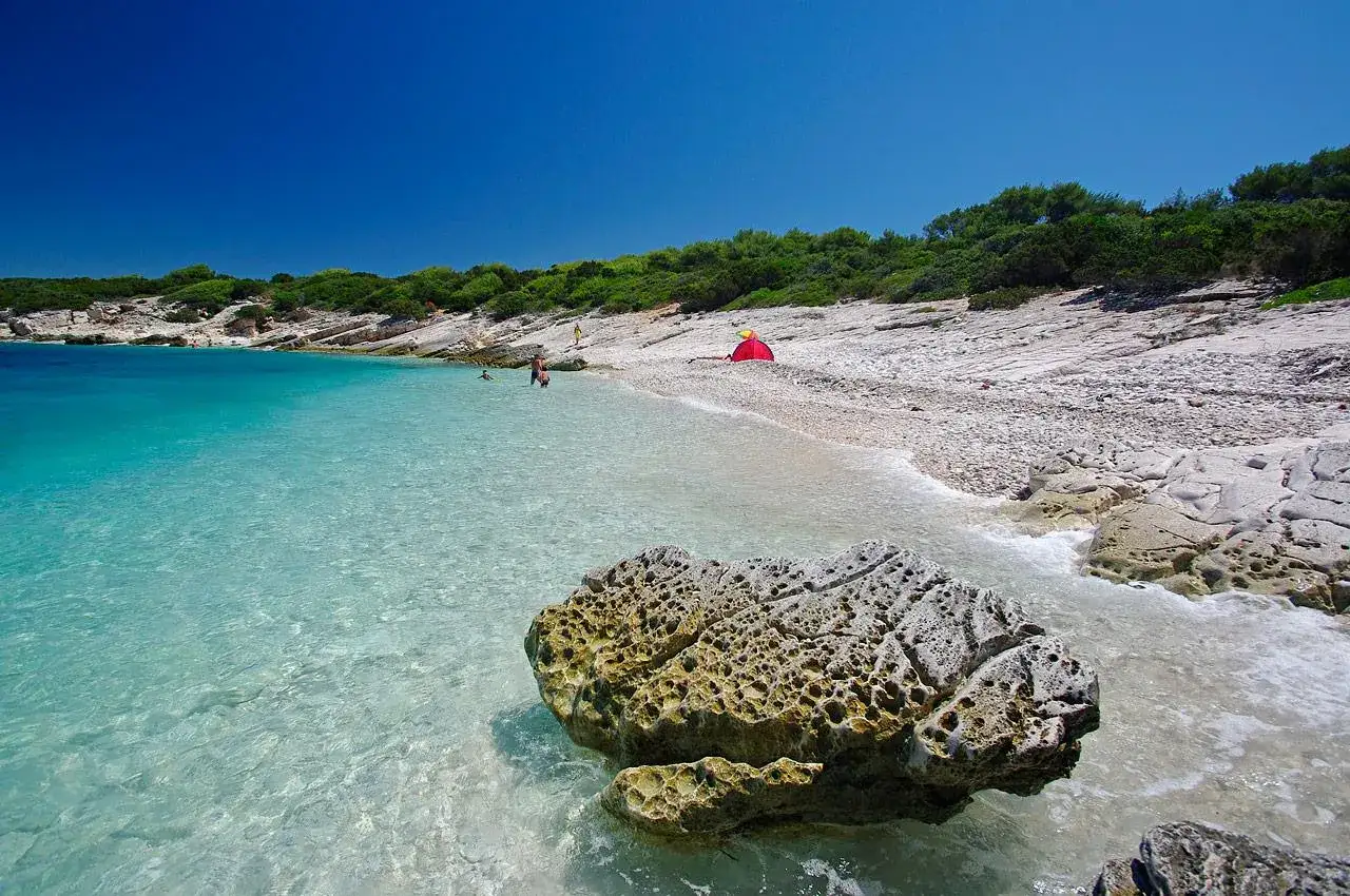 Piękna, skalista wyspa Proizd z turkusową wodą i kamienistą plażą. Czerwony namiot i kilka osób odpoczywa w słońcu.