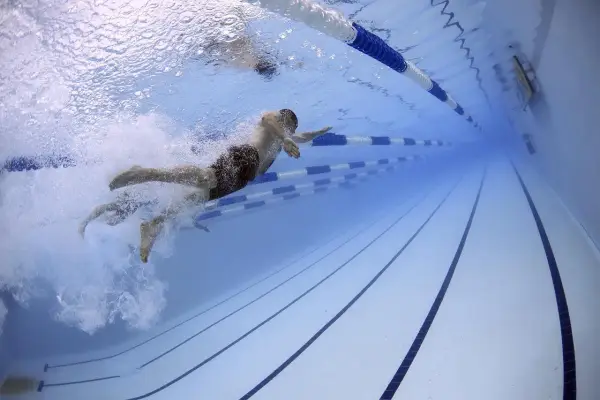 Pływak w ciemnych kąpielówkach pokonuje tor w basenie. Woda jest przejrzysta, a światło odbija się od powierzchni. To pokazuje, jak utrzymać się na wodzie dzięki technice.