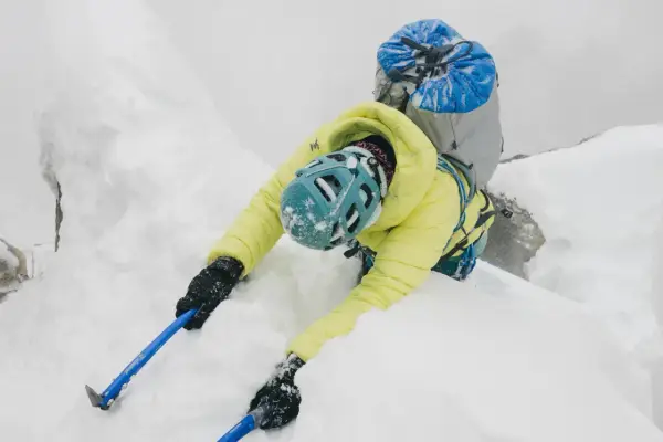 Alpinista w żółtej kurtce i niebieskim kasku wspina się po śnieżnej grani, używając czekana do hamowania.