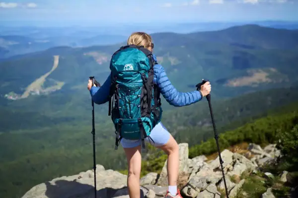 Kobieta z turkusowym plecakiem w góry, z kijkami trekkingowymi, podziwia panoramę.