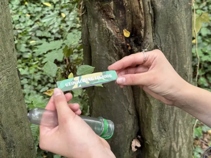 Ręce trzymają pojemnik i zeszyt z napisem "GEOCACHING LOGBOOK". To wyjaśnia, geocaching co to jest: poszukiwanie ukrytych skarbów.
